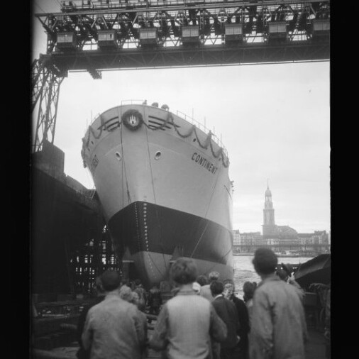 Bugansicht des Schiffs CONTINENT auf einer Werft-Slipanlage während des Stapellaufs, mehrere Zuschauer im Vordergrund, ein hohes Portalkran-Gerüst mit Schriftzug darüber und ein Kirchturm am gegenüberliegenden Ufer im Hintergrund.