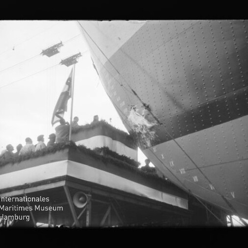 Schwarz-weiß-Aufnahme: Menschen auf einem geschmückten Tribünenpodium mit Flagge beobachten ein großes Luftschiff, dessen Außenhülle rechts deutlich einen Riss/Loch und Nieten zeigt.
