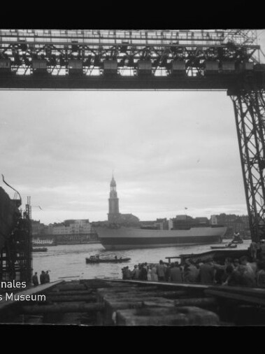 Schwarzweißfoto, durch das Stahlgerüst eines großen Auslegerkrans gerahmter Blick auf ein großes Schiff im Hafen, vor dem Ufer eine Menschenmenge und im Hintergrund eine Stadtsilhouette mit hohem Kirchturm; linke untere Ecke zeigt das Logo des Internationalen Maritimen Museums Hamburg.