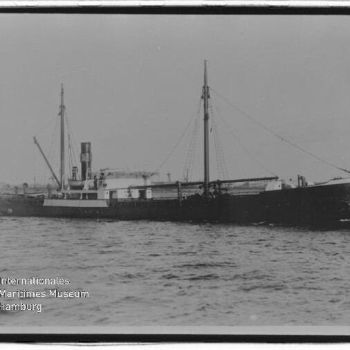Schwarzweißaufnahme eines historischen Frachtschiffs in Seitenansicht auf ruhiger See, mit prominentem Schornstein, zwei Masten und Ladebaum; unten links sichtbar das Logo des Internationalen Maritimen Museums Hamburg.