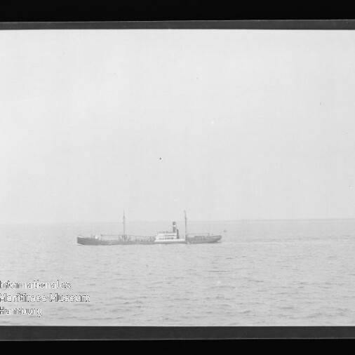 Schwarzweiß-Foto eines kleinen Frachtdampfers mittig am Horizont auf ruhiger See unter weitem Himmel, im unteren linken Bildbereich ein Wasserzeichen des Internationalen Maritimen Museums Hamburg.