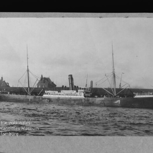 Schwarz-weiß-Fotografie eines großen Dampfschiffs mit zentralem Schornstein und zwei Masten sowie Ladebäumen, längsseits am Kai vor historischen Gebäuden, welliges Wasser im Vordergrund und Museumswasserzeichen unten links.