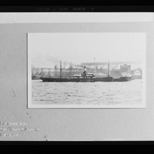 Gerahmtes Schwarzweißfoto eines dampfgetriebenen Frachtschiffs auf dem Wasser vor einer Industriehafen-Kulisse, auf hellem Karton montiert und mit dem Stempel 'Internationales Maritimes Museum Hamburg' unten links.