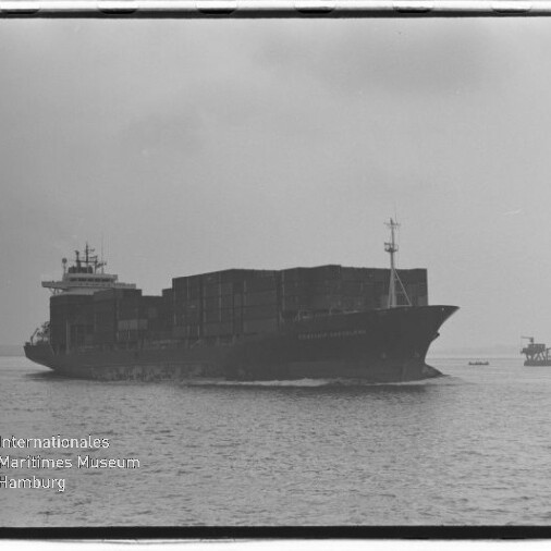 Schwarzweißaufnahme eines beladenen Containerschiffs mit gestapelten Containerkästen, das bei ruhiger See vor bedecktem Himmel fährt; rechts im Hintergrund eine kleinere Plattform und links unten das Logo des Internationalen Maritimen Museums Hamburg.