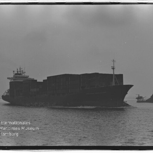 Schwarz-weiß-Foto eines großen, beladenen Containerschiffs, das bei ruhigem Wasser und bedecktem Himmel fährt und rechts von einem kleinen Schlepper begleitet wird; unten links sichtbares Wasserzeichen des Internationalen Maritimen Museums Hamburg.