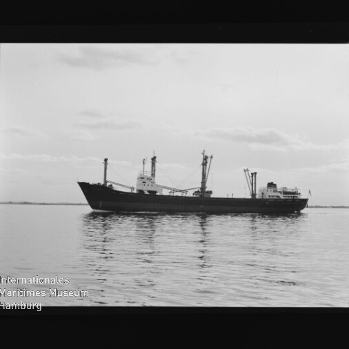 Schwarzweißfoto eines Frachtschiffs in Seitenansicht auf ruhiger See unter leicht bewölktem Himmel, mit Museumswasserzeichen unten links.