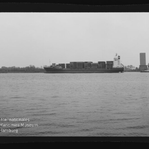 Schwarzweiß-Foto eines großen Containerschiffs mit gestapelten Ladecontainern, das auf ruhigem Wasser vor einer flachen Uferlinie mit Bäumen und einem hohen Lager- oder Bürogebäude fährt; Wasserzeichen des Internationalen Maritimen Museums Hamburg unten links.