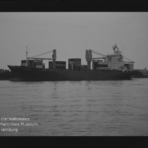 Schwarzweißaufnahme eines Frachtschiffs mit aufgestapelten Containern und zwei Ladekränen, das auf einem ruhigen Fluss vorbeifährt; unten links sichtbar das Wasserzeichen des Internationalen Maritimen Museums Hamburg.