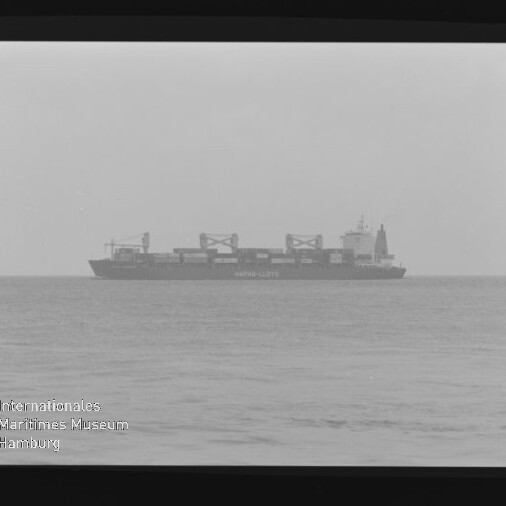 Containerfrachter mit mehreren Deckskränen auf ruhiger See am Horizont unter nebligem Himmel, das Foto im Filmrahmen zeigt unten links das Wasserzeichen des Internationalen Maritimen Museums Hamburg.