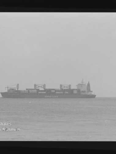 Schwarzweißaufnahme eines Containerschiffs auf ruhigem Meer am Horizont, deutlich sichtbar Containerstapel und Ladekräne, mit Filmrand und Wasserzeichen des Internationalen Maritimen Museums Hamburg.