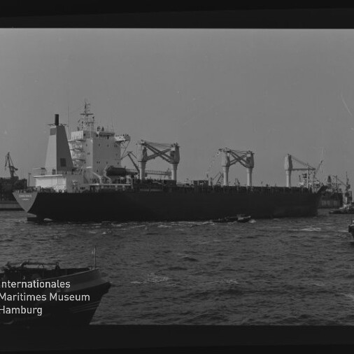 Schwarzweißaufnahme eines seitlich sichtbaren Frachtschiffs mit mehreren Deckkränen, das durch einen Hafen fährt; im Vordergrund schäumendes Wasser und kleine Boote, im Hintergrund Hafenkräne und das Logo des Internationalen Maritimen Museums Hamburg.