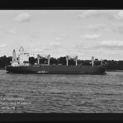 Schwarzweißaufnahme eines Massengutfrachters mit drei Ladekränen, daneben ein kleines Lotsenboot, ruhiges Wasser und baumgesäumte Küste unter bewölktem Himmel; Internationales Maritimes Museum Hamburg‑Logo links unten.