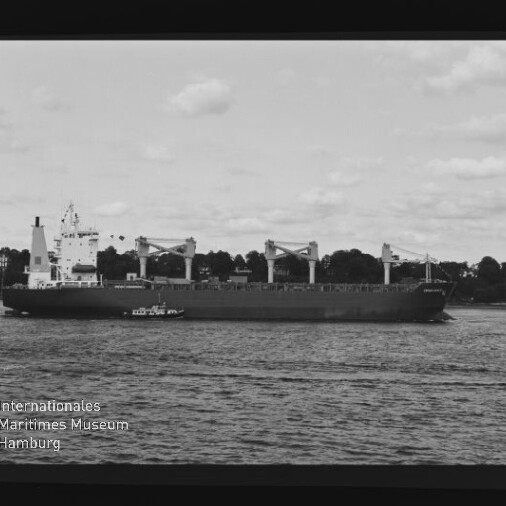Schwarzweiß-Foto eines großen Frachtschiffs mit vier Ladekränen, begleitet von einem Lotsenboot auf dem Wasser vor einer baumbestandenen Uferlinie; sichtbarer Filmrand und Logo des Internationalen Maritimen Museums Hamburg.