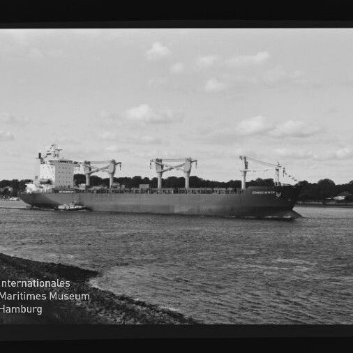 Schwarzweißaufnahme eines großen Frachtschiffs mit mehreren Deckskranen, begleitet von einem Schlepper, das nahe der Uferlinie vor baumbestandener Küste fährt; unten links das Logo des Internationalen Maritimen Museums Hamburg.