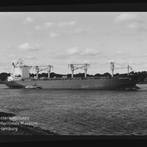 Schwarzweiß-Foto eines großen Frachtschiffs mit drei Deckskränen und einem kleinen Schlepper an seiner Seite, das auf ruhigem Wasser vor einer baumbewachsenen Küstenlinie fährt; Filmrandmarkierungen und Museumswasserzeichen sind im Bild sichtbar.
