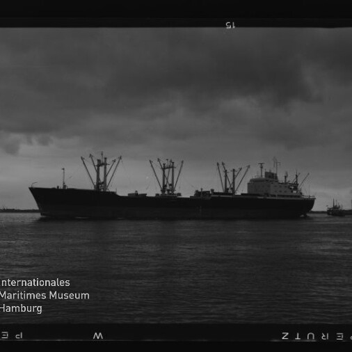 Schwarzweiß-Foto eines großen Frachtschiffs mit aufragenden Ladekränen, das vor düsterem, wolkenverhangenem Himmel auf ruhigem Wasser liegt; rechts ein kleiner Schlepper und links unten das Logo des Internationalen Maritimen Museums Hamburg sowie sichtbarer Filmrand.