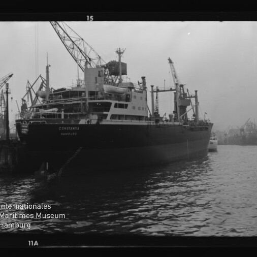 Schwarzweiß-Foto eines großen Frachtschiffs mit dem Namen „Constantia“, das am Kai eines Industriehafens liegt, umgeben von Hafenkränen und weiteren Schiffen im dunstigen Hintergrund.