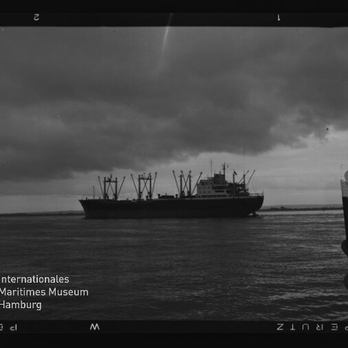 Schwarzweiß-Fotografie eines Frachtschiffs mit mehreren Ladekränen auf ruhigem Wasser unter dunklen Wolken; rechts im Vordergrund ist der Bug eines weiteren Bootes und unten links das Logo des Internationalen Maritimen Museums Hamburg zu sehen.