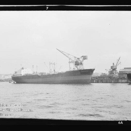 Schwarzweiß-Foto eines großen Frachtschiffs, das am Kai eines Industriehafens mit mehreren Ladekränen und Hafenbauten liegt; im Vordergrund Wasser, sichtbar sind Filmränder und ein Wasserzeichen des Internationalen Maritimen Museums Hamburg.