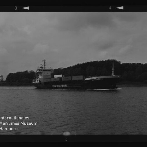 Schwarzweißaufnahme eines Containerschiffs mit der Aufschrift CONTAINERSHIPS, das auf einem ruhigen Fluss oder Kanal entlangfährt, vor bewaldetem Ufer und bewölktem Himmel; Museumslogo unten links.