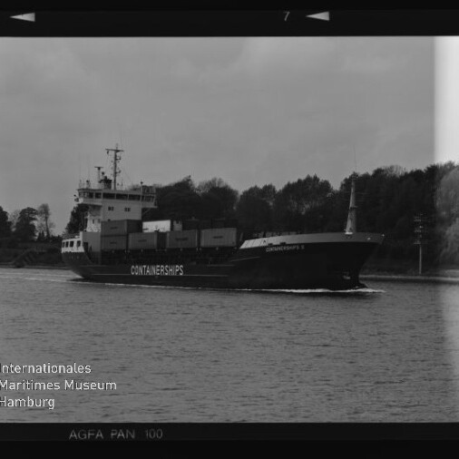 Schwarz-weiß-Foto eines Containerschiffs mit dem Schriftzug CONTAINERSHIPS, beladen mit Containern und fahrend auf ruhigem Wasser vor einer baumbestandenen Uferlinie; Filmkanten und das Logo des Internationalen Maritimen Museums Hamburg sind im Bildrand sichtbar.