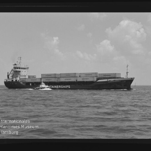 Schwarzweiß-Foto eines beladenen Containerschiffs mit Aufschrift CONTAINERSHIPS, neben dem ein kleines Lotsenboot fährt; ruhige See, vereinzelte Wolken am Horizont sowie sichtbarer Filmrand und Wasserzeichen des Internationalen Maritimen Museums Hamburg