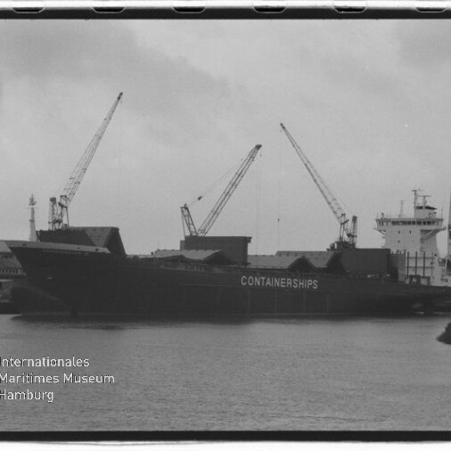 Schwarzweißaufnahme eines großen Containerschiffs mit der Aufschrift „CONTAINERSHIPS“, festgemacht am Kai neben mehreren Hafenkränen, Wasserfläche im Vordergrund und Museumslogo unten links.