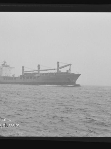 Schwarzweißaufnahme eines Frachtschiffs mit drei Ladekränen auf offenem, leicht nebligem Meer; bewegte See im Vordergrund und unten links das Wasserzeichen des Internationalen Maritimen Museums Hamburg.