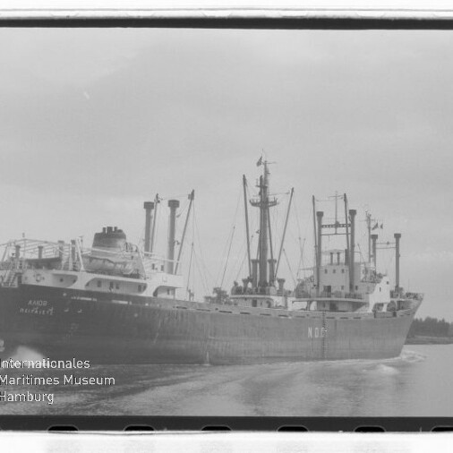 Schwarzweißfoto eines großen Frachtschiffs mit mehreren Masten und Ladekränen, das auf einem Kanal fährt und eine Bugwelle zieht; links unten Wasserzeichen des Internationalen Maritimen Museums Hamburg.