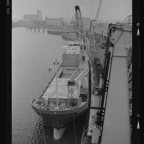 Schwarzweißaufnahme eines beladenen Frachtschiffs am Industrie-Kai mit mehreren Hafenkränen und Lagergebäuden im Nebel im Hintergrund, unten links Logo des Internationalen Maritimen Museums Hamburg.