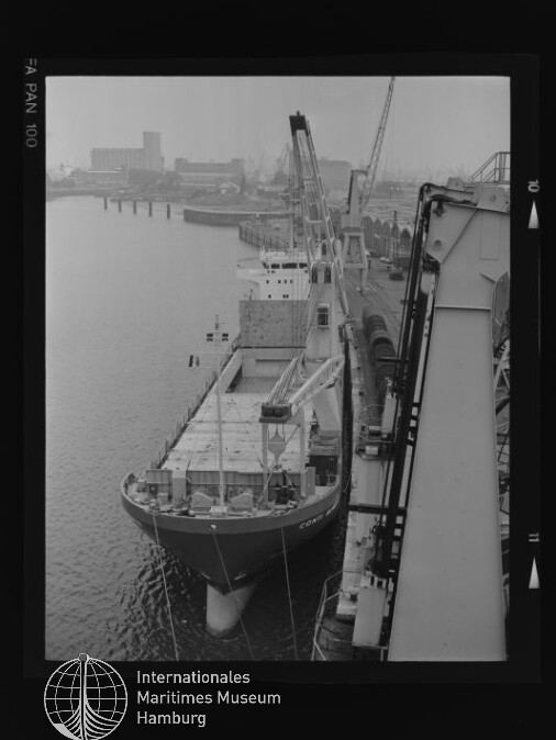 Schwarzweißaufnahme eines beladenen Frachtschiffs am Industrie-Kai mit mehreren Hafenkränen und Lagergebäuden im Nebel im Hintergrund, unten links Logo des Internationalen Maritimen Museums Hamburg.