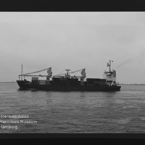 Schwarzweißaufnahme eines Frachtschiffs mit gestapelten Containern und zwei Ladekränen, das auf ruhiger See vor flachem, wolkenverhangenem Horizont fährt; Filmrand und das Logo des Internationalen Maritimen Museums Hamburg sind sichtbar.