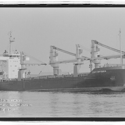 Schwarzweiß-Foto eines Frachtschiffs mit drei sichtbaren Ladekränen und dem Namen CONTI BRITANNIA am Bug, das ruhig im Wasser vor einem Hafen liegt; links unten Wasserzeichen des Internationalen Maritimen Museums Hamburg.