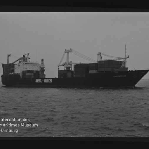 Schwarzweißaufnahme eines fahrenden Containerschiffs mit mehreren Deckskränen und gestapelten Containern auf ruhigem Meer, flachem Horizont und Museumswasserzeichen unten links.