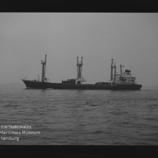 Schwarzweißaufnahme eines Frachtschiffs mit drei hohen Ladekränen, das auf ruhiger See vor flachem Horizont fährt; sichtbar sind Filmrand mit der Aufschrift „SAFETY FILM“ und ein Museumswasserzeichen.