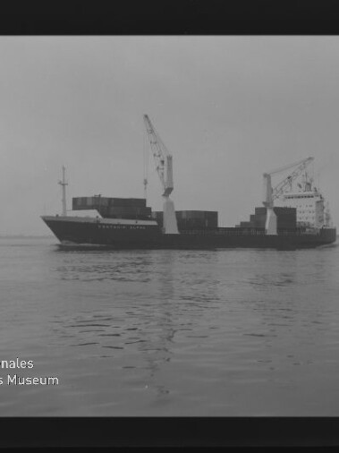 Schwarzweiß-Foto eines Frachtschiffs mit zwei Ladekränen und gestapelten Containern auf ruhigem Wasser, links teilweise ein zweites Schiff sichtbar und unten links das Logo des Internationalen Maritimen Museums Hamburg.