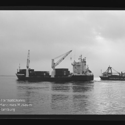 Schwarzweißaufnahme zweier Frachtschiffe auf ruhigem Wasser: links ein containerbeladener Frachter mit Ladekränen, rechts ein kleineres Schiff mit Kran und der Aufschrift ‚Hamburg-Linien‘, vor bedecktem Himmel und mit sichtbarem Museumslogo und Filmrand.