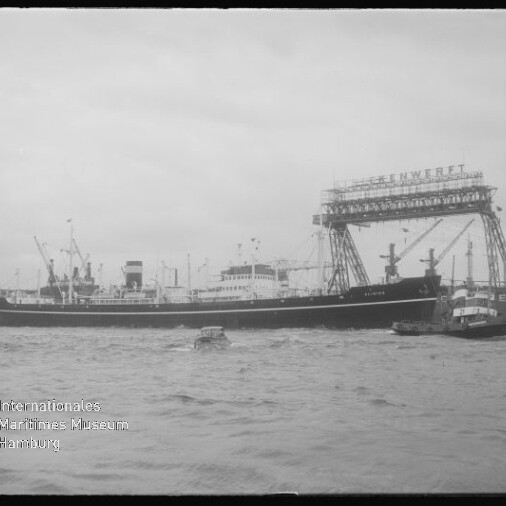 Schwarzweißaufnahme eines großen Frachtschiffs im Hafen vor einer Werft mit massivem Portalkran und mehreren Kränen, begleitet von kleineren Begleitbooten auf dem Wasser.