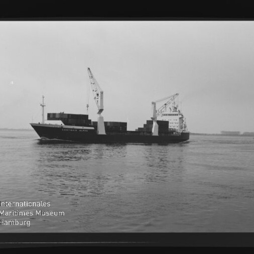 Schwarzweißaufnahme eines Frachtschiffs mit Containern und zwei Ladekränen, das ruhig auf dem Meer vor einer flachen Küstenlinie fährt; unten links das Logo des Internationalen Maritimen Museums Hamburg sichtbar.