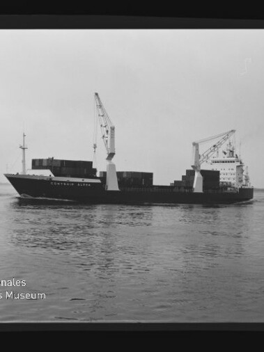 Schwarzweißaufnahme eines Containerfrachters mit zwei Bordkränen und gestapelten Containern, fahrend auf ruhigem Wasser; links ein weiteres Schiff am Bildrand und unten links das Logo des Internationalen Maritimen Museums Hamburg, sichtbar innerhalb des Filmrands.
