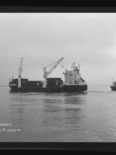 Schwarzweiß-Aufnahme eines Frachtschiffs mit ausfahrbaren Ladekränen und Containerstapel, rechts ein weiteres Schiff mit dem Schriftzug HAMBURGLINJEN, auf ruhiger See unter trübem Himmel; unten links Logo und Schriftzug des Internationalen Maritimen Museums Hamburg.