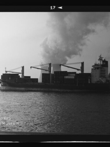 Schwarzweißaufnahme eines Containerfrachters mit Ladekränen auf ruhigem Wasser, vor einer großen Rauch- oder Dampfwolke und neben einer hohen Strommastkonstruktion; Wasserzeichen des Internationalen Maritimen Museums Hamburg unten links.