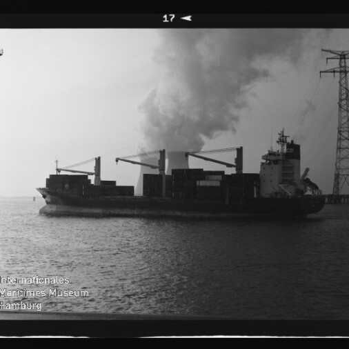 Schwarzweißaufnahme eines Containerfrachters mit Ladekränen auf ruhigem Wasser, vor einer großen Rauch- oder Dampfwolke und neben einer hohen Strommastkonstruktion; Wasserzeichen des Internationalen Maritimen Museums Hamburg unten links.