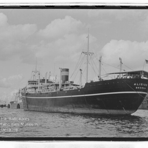 Großes Frachtschiff „Alisios Bremen“ längsseits im Hafen, Seitenansicht mit markantem Schornstein, Ladekranen und Flagge am Heck vor bewölktem Himmel.