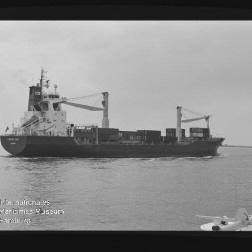 Schwarzweiß-Seitenansicht eines Frachtschiffs mit gestapelten Containern und zwei Ladekränen, das auf ruhigem Wasser fährt; im Vordergrund rechts ein Teil eines Wasserflugzeugs und unten links das Logo des Internationalen Maritimen Museums Hamburg sichtbar.