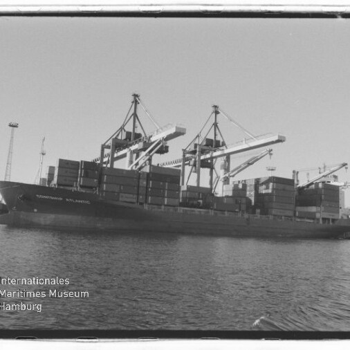 Schwarzweiß-Foto eines mit Containern beladenen Frachtschiffs am Hafenpier, davor Wasser und mehrere große Containerkräne sowie Hafeninfrastruktur; Wasserzeichen des Internationalen Maritimen Museums Hamburg in der Ecke.