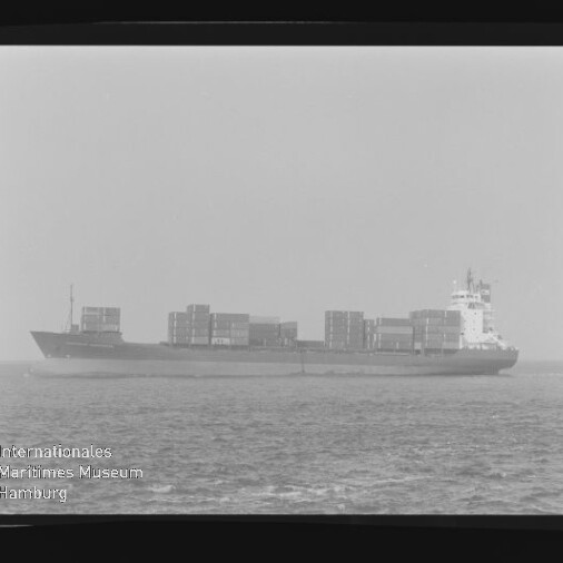 Schwarz-weiß-Foto eines Containerschiffs in Seitenansicht auf ruhiger See mit übereinandergestapelten Ladecontainern, leicht diesigem Horizont, sichtbarem Filmrand und der Aufschrift AGFA APX 100 sowie Museumslogo unten links
