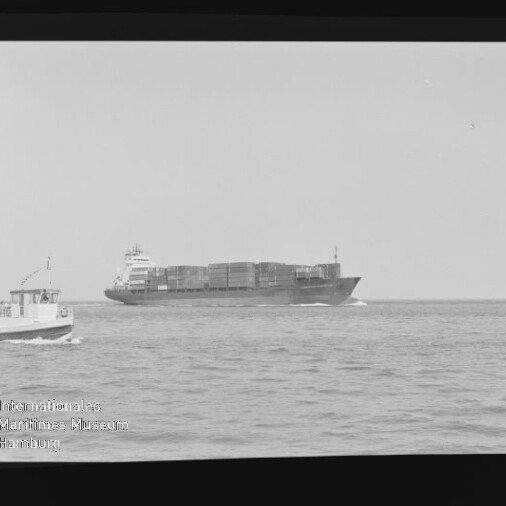 Schwarzweißaufnahme eines großen Containerschiffs auf offenem Meer, links vorn ein kleines Ausflugsboot, umgeben von Filmrand mit Schriftzug AGFA APX 100 und Museumslogo.