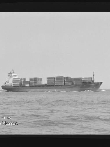 Schwarzweiß-Foto eines beladenen Containerschiffs in Seitenansicht mit aufgetürmten Containerblöcken, das auf ruhiger See fährt und Bug- sowie Heckwellen zeigt; schwarzer Filmrand mit Nummerierung und Wasserzeichen des Internationalen Maritimen Museums Hamburg sind sichtbar.
