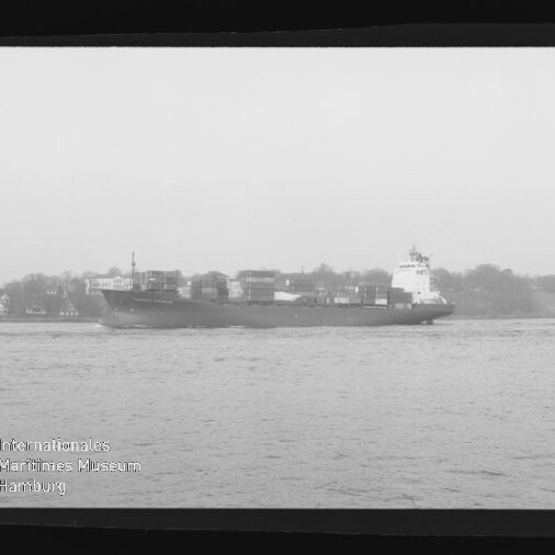 Schwarzweiß-Aufnahme eines Containerschiffs mit aufgestapelten Containern, das auf ruhigem Wasser vor einem Ufer mit Gebäuden und Bäumen fährt; schwarzer Filmrand und Museumslogo unten links sichtbar.